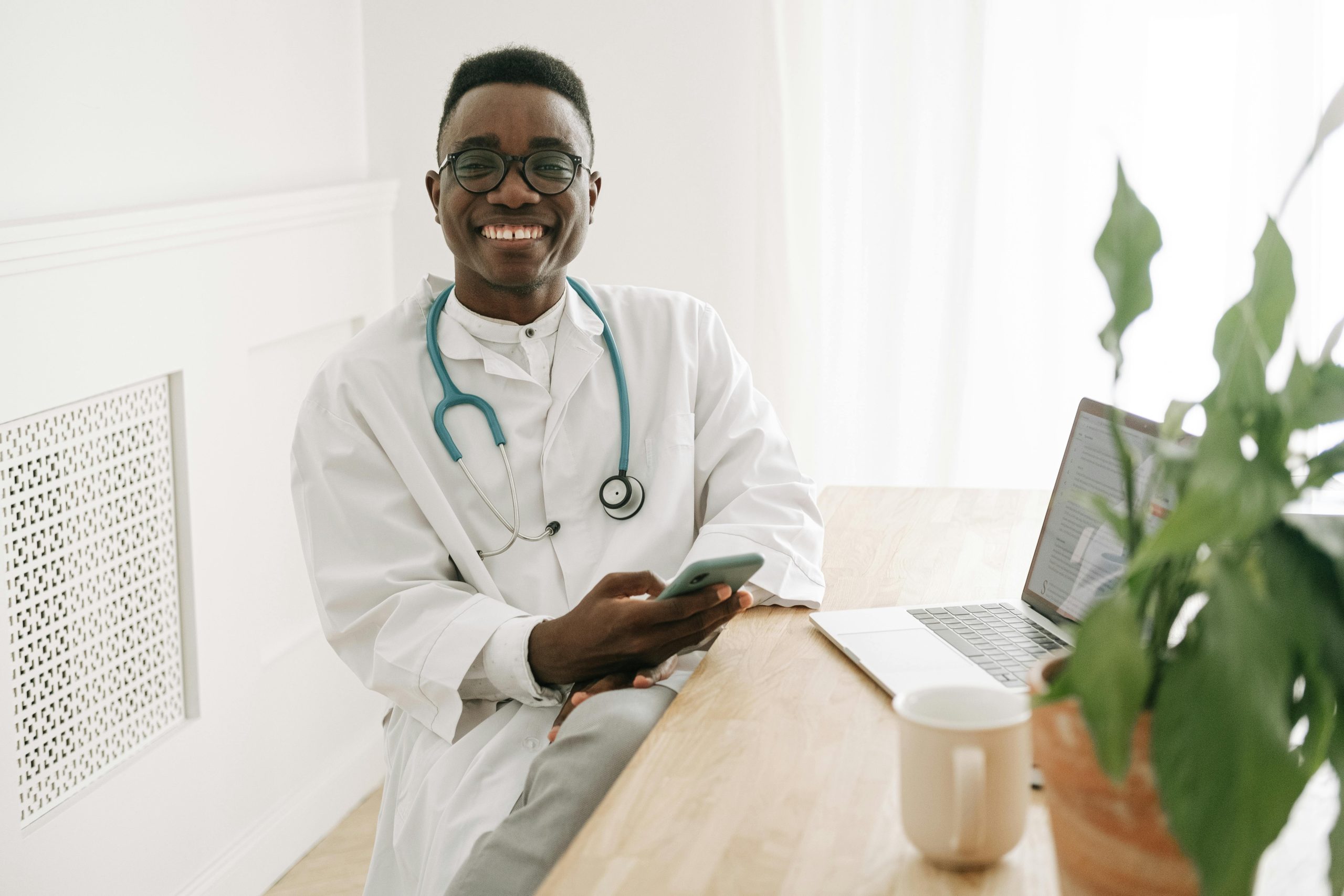 project management for healthtech companies A man in scrubs with a stethoscope in front of a computer, holding a phone, and smiling at the camera.