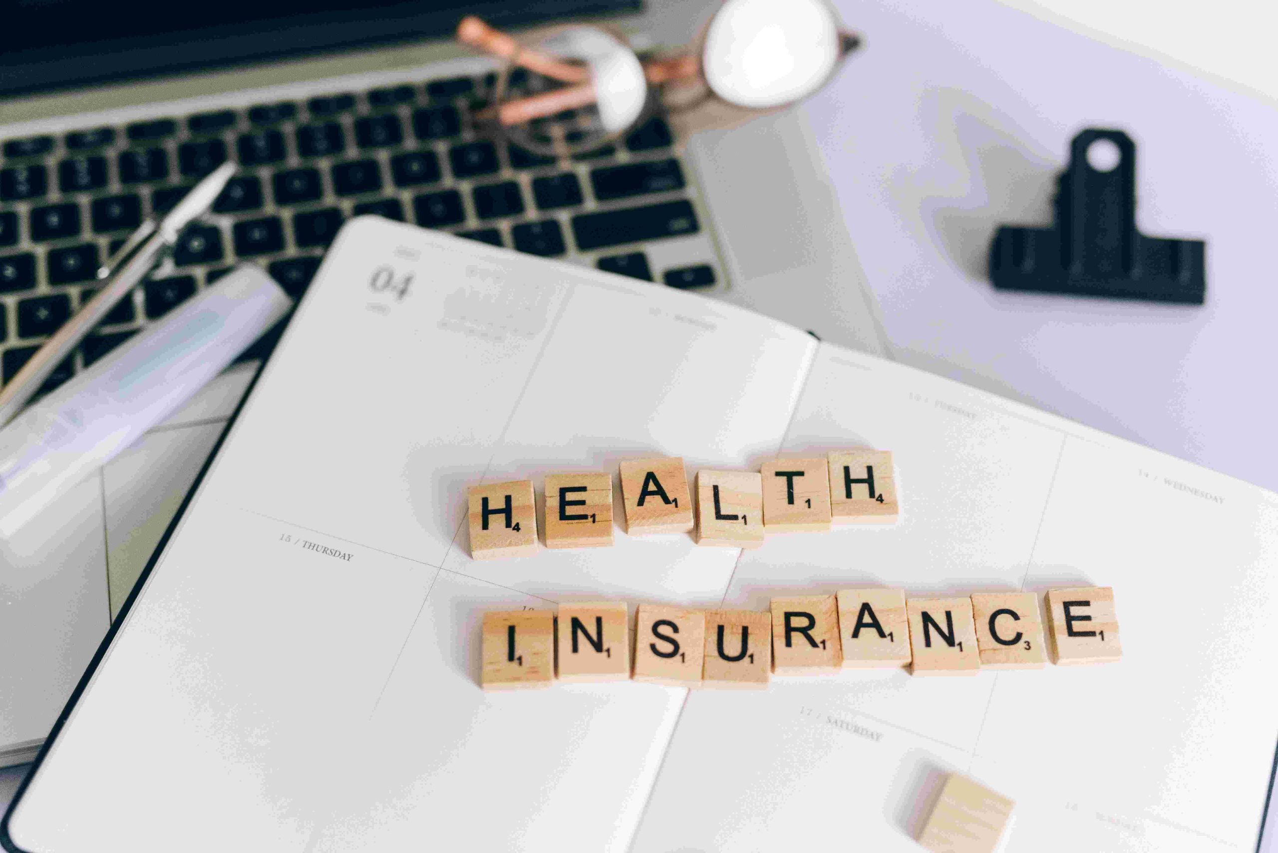 Scrabble tiles spelling out "Health Insurance" against a messy desk background.